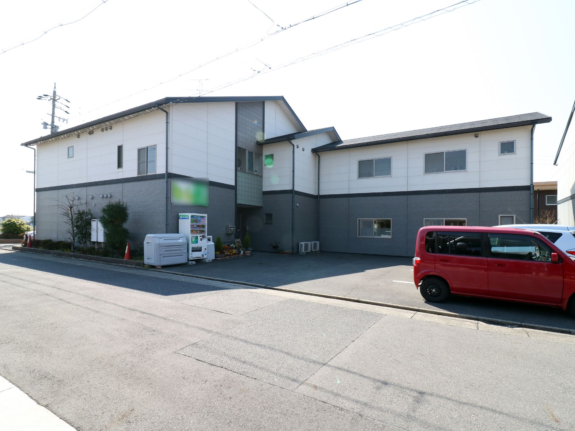 施設は白とグレーの三角屋根の2階建ての建物。通りに面していて駐車場があり、乗用車が複数台駐車されている。入り口前には自動販売機もある。