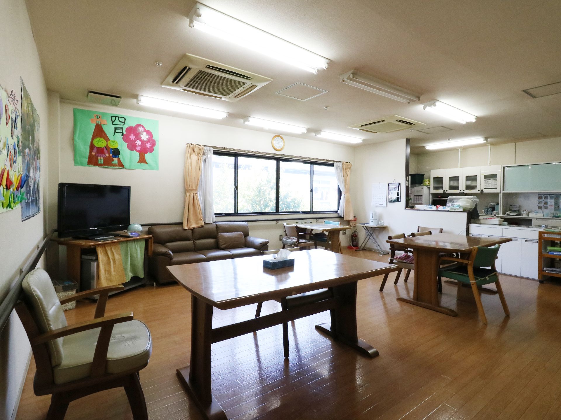 施設の食堂は白壁にフローリングの床。奥には横長の窓がありカーテンもある。天井にはエアコンがあり奥にはキッチンが見える。