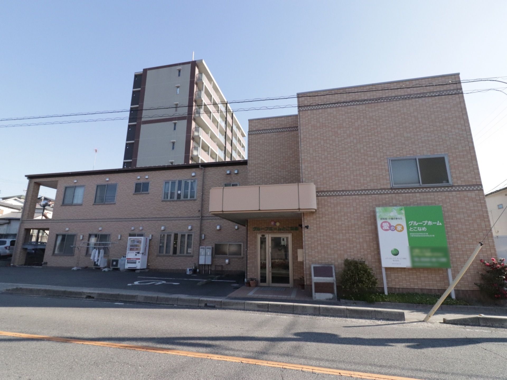 広い道路に面している2階建ての建物でエントランス横には車椅子専用の駐車場や自販機、物干しざおがある。