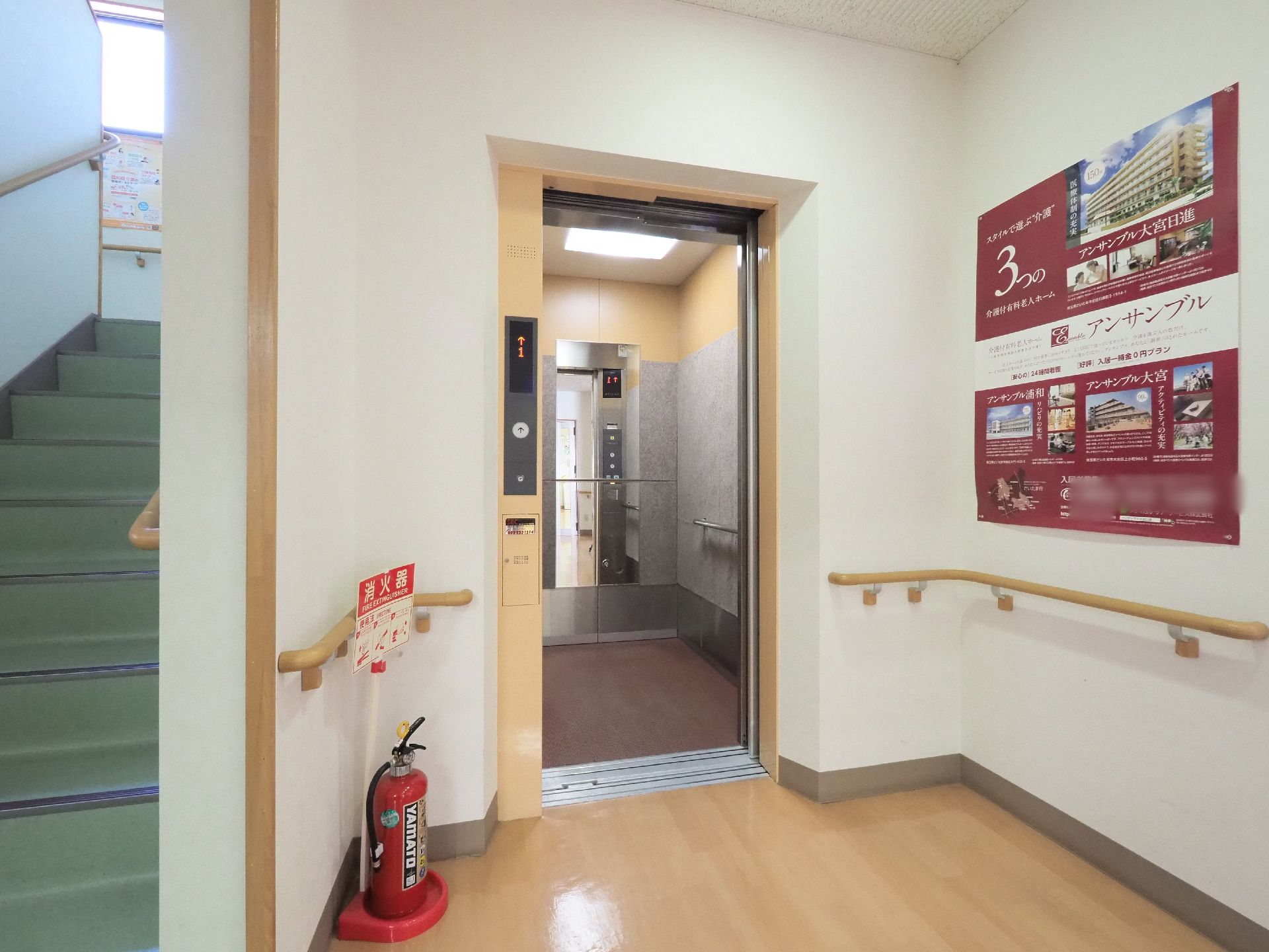 施設の階段の横には茶色のエレベーターが設置されている。奥行きがあり鏡や手すりもある。近くには消火器も置かれている。