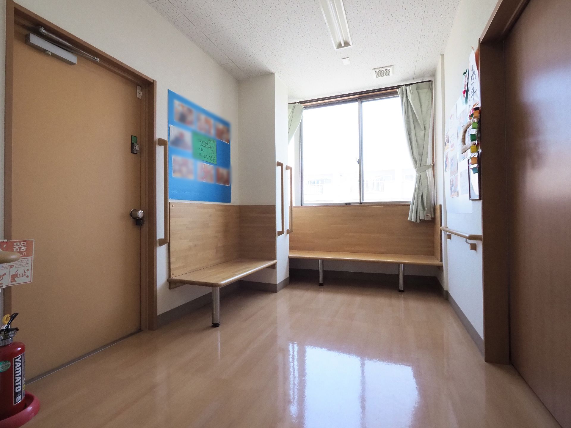 廊下の突き当りに備え付けのベンチが2つある。窓があり、カーテンがつけてある。壁に模造紙が貼ってあり、写真が飾ってある。