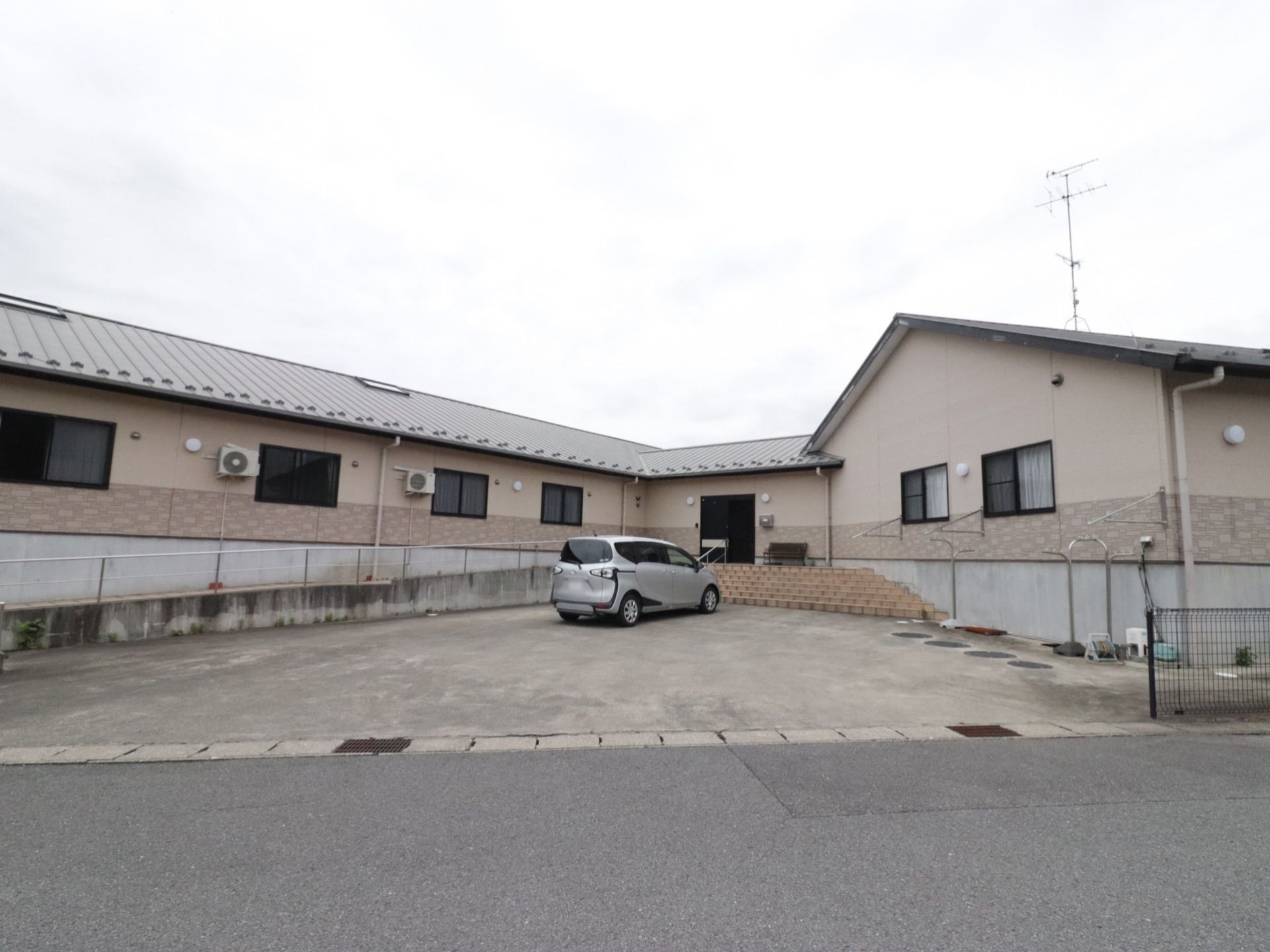 ブラウンの外壁の建物で、通常の住宅のような雰囲気である。建物の前には駐車スペースがあり、車が１台止まっている。