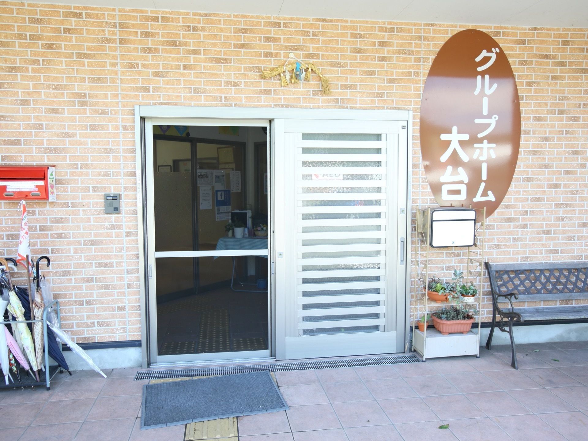 中央に銀色の引き戸の玄関扉があり、扉の横には大きくホームの名称が書かれた長円形のプレートが貼られている。