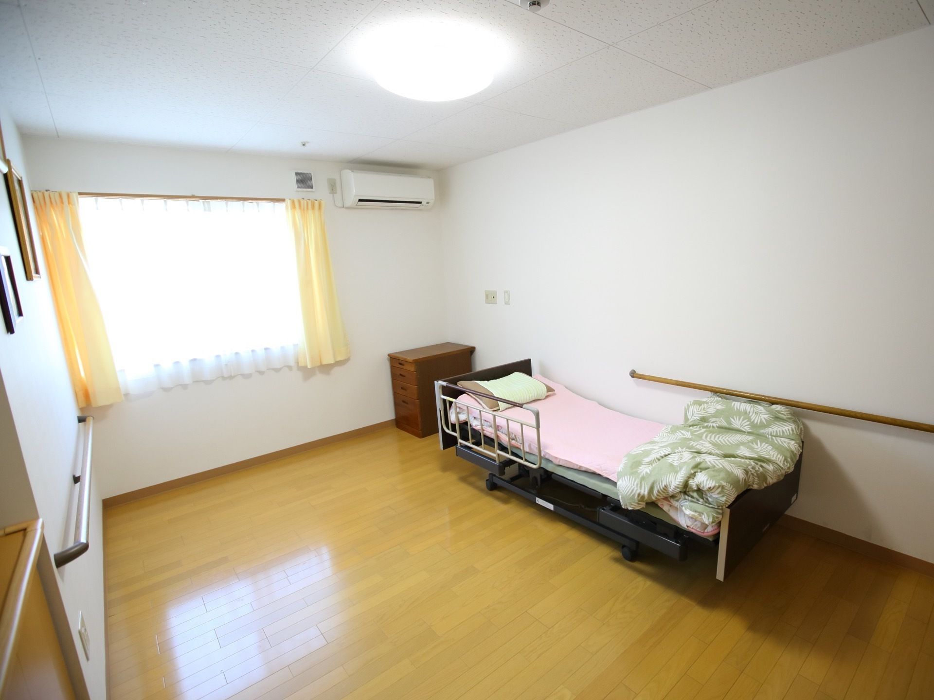 エアコンが備え付けられた居室。介護用のベッドとタンスなどが室内に置かれており、窓からは陽の光が差し込んでいる。