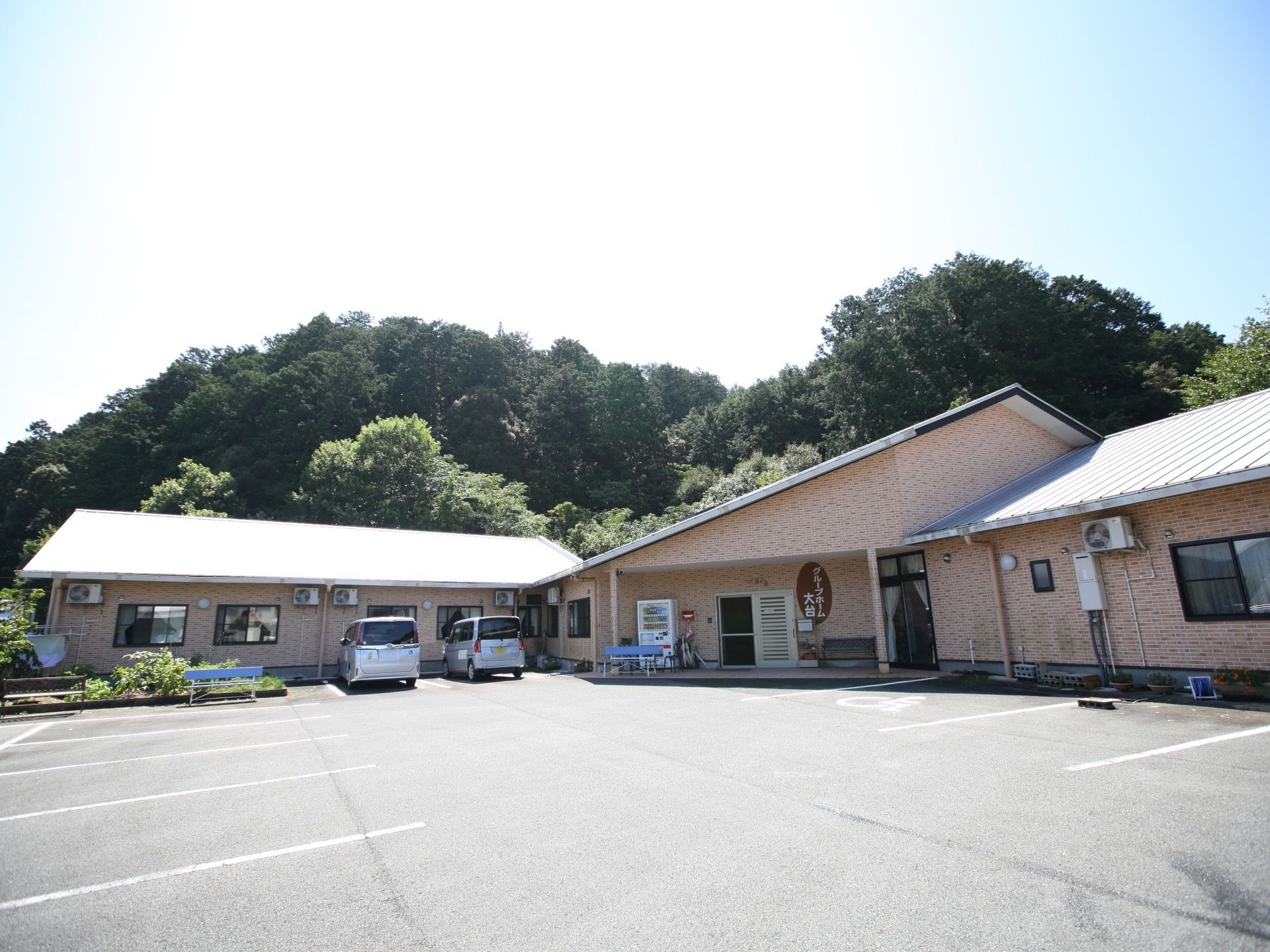 暖色系の外壁をした平屋建てのホーム。玄関の横には大きく施設名が書かれたプレートが掲示されている。ホームの前面には駐車場と畑がある。