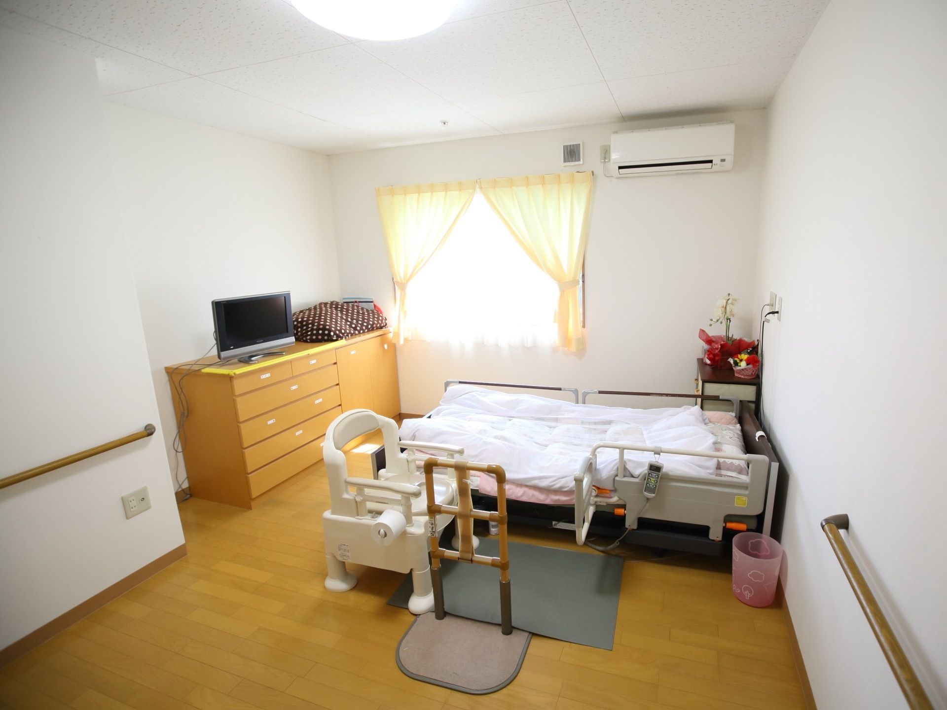 エアコンと介護用ベッド、簡易トイレ、収納家具などが備え付けられた居室。壁の一部には手すりも設置されている。