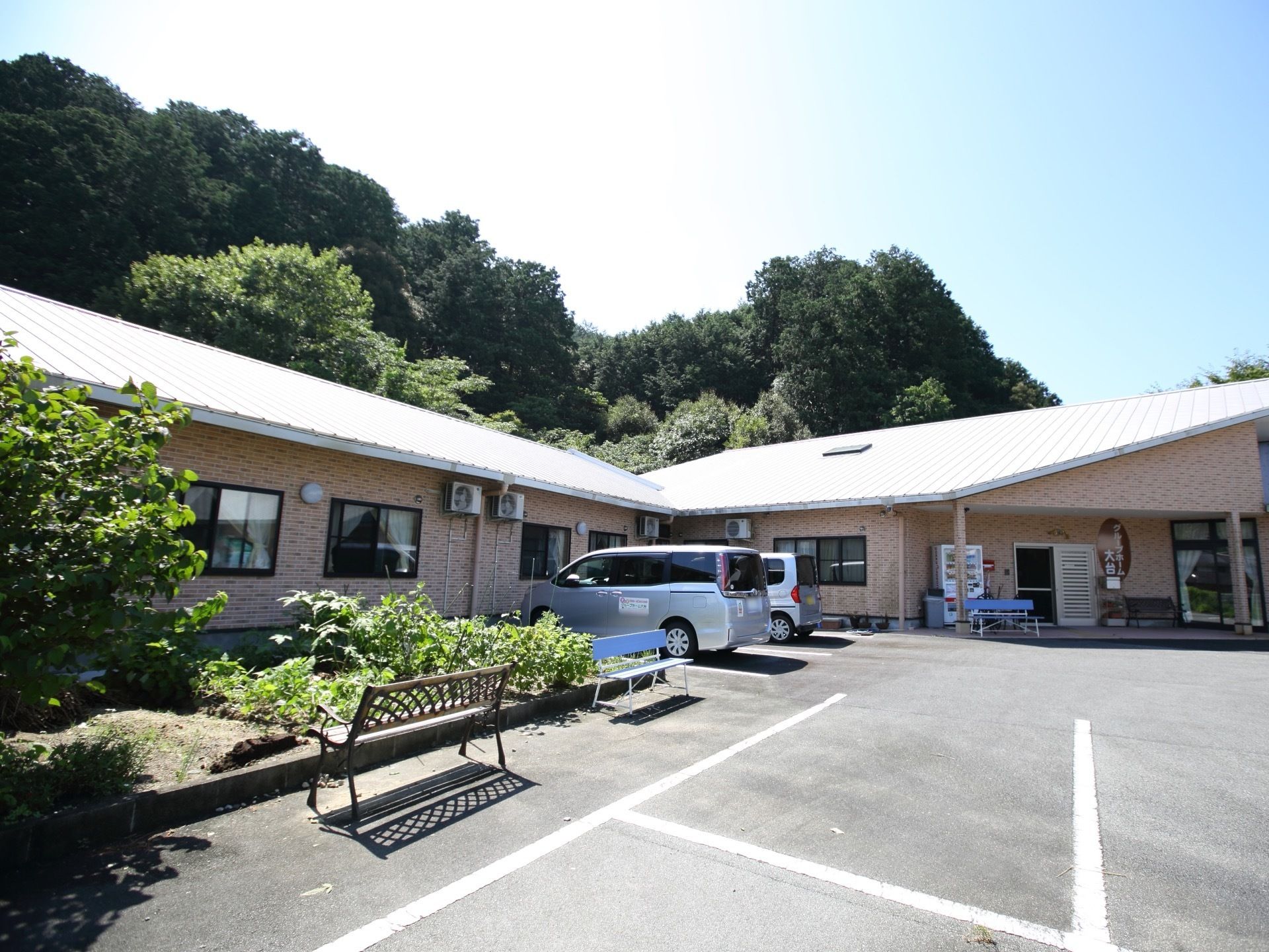 平屋建ての大きなホーム。ホームの前には、畑と駐車場があり、駐車場にはホームの送迎車などが停車しているのが見える。