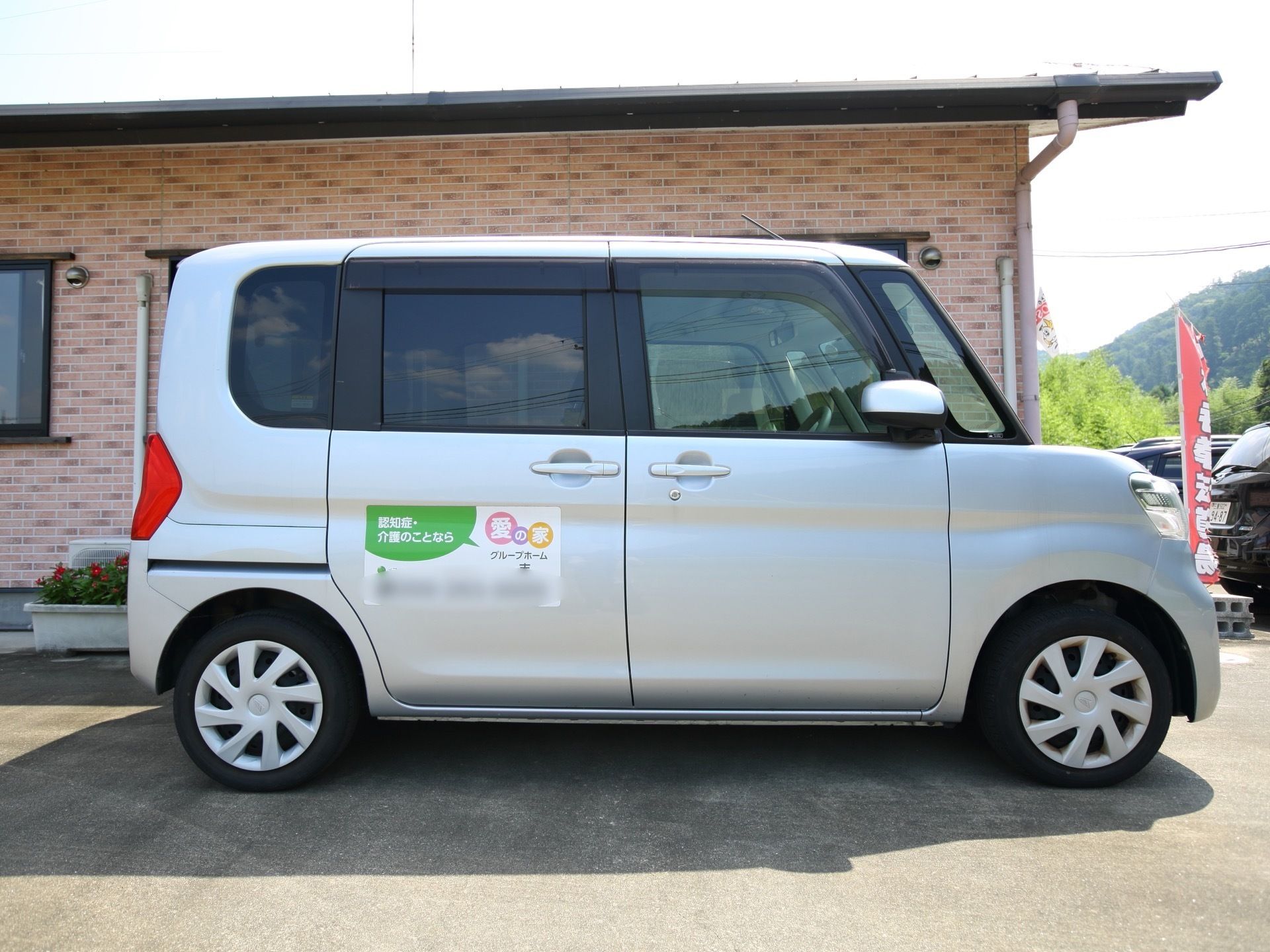 シルバーの施設車がホームの隣に駐車されており、後部座席は乗り降りのしやすいスライド式のドアである。後部ドアには施設名が表示されている。