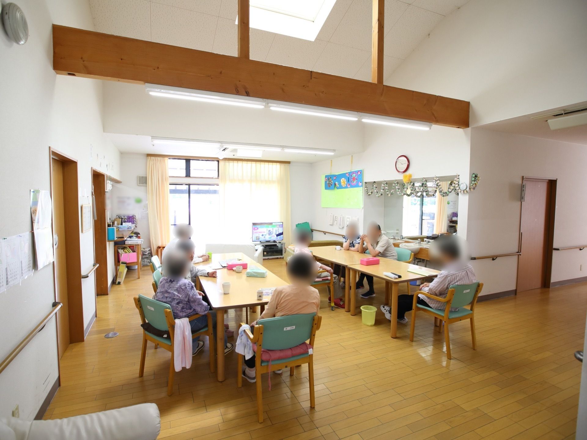 共有スペースでは入居者様がお茶を飲みながらテレビ鑑賞や談笑を楽しんでいる。大きな窓があり明るく開放的である。