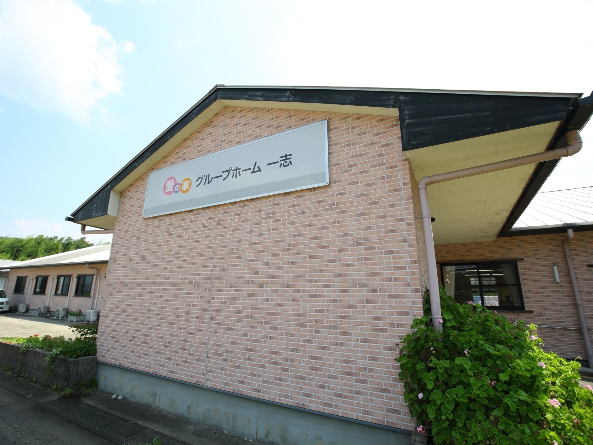 淡いピンク色をした平屋造りの建造物である。シンプルなデザインの施設看板が壁に取り付けられている。日当たりが良く、後方には木々が見える。