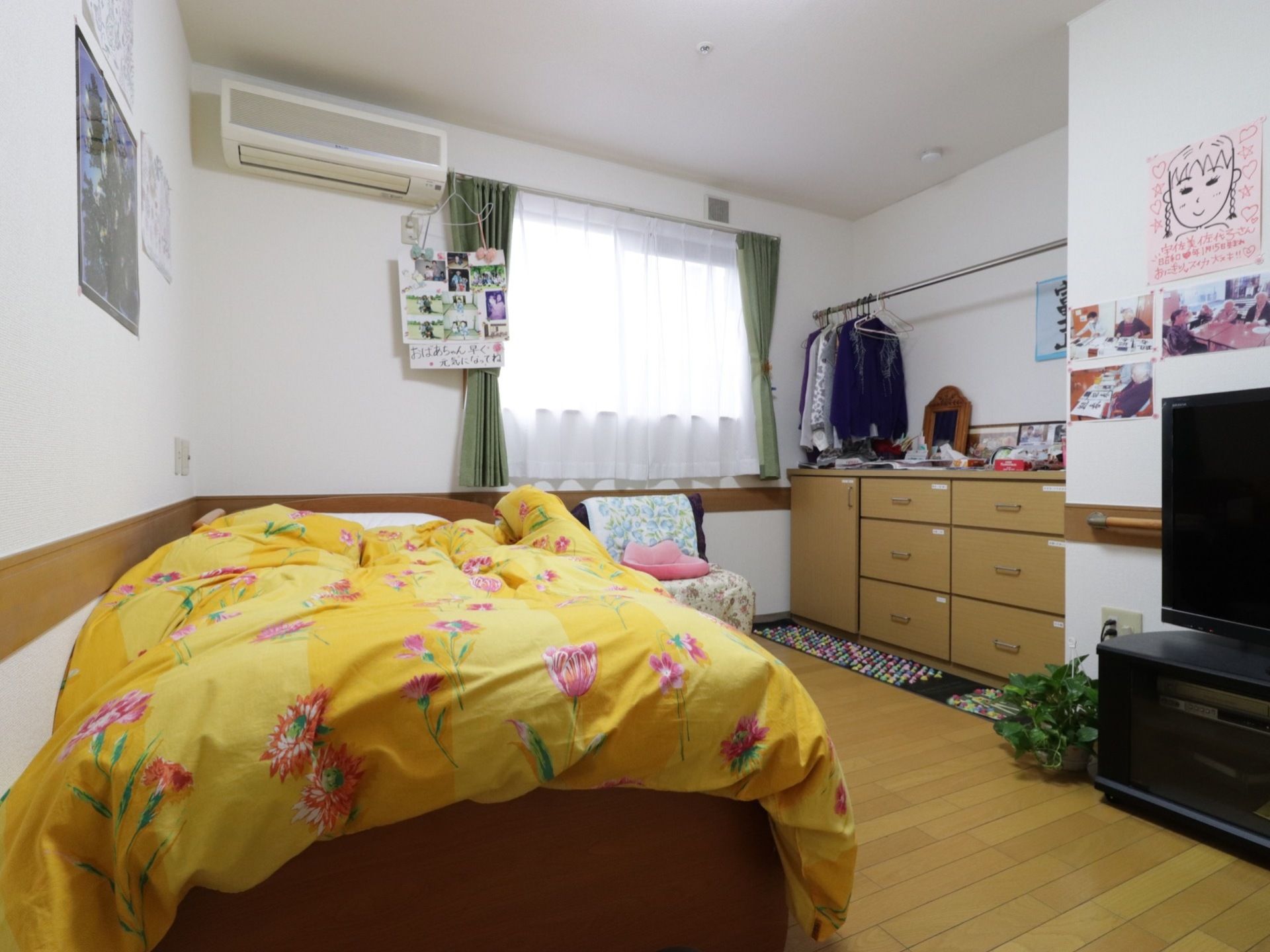 居室には黄色の布団があるベッドをはじめ、テレビやタンス、洋服をかけるバーがある。壁には似顔絵もある。