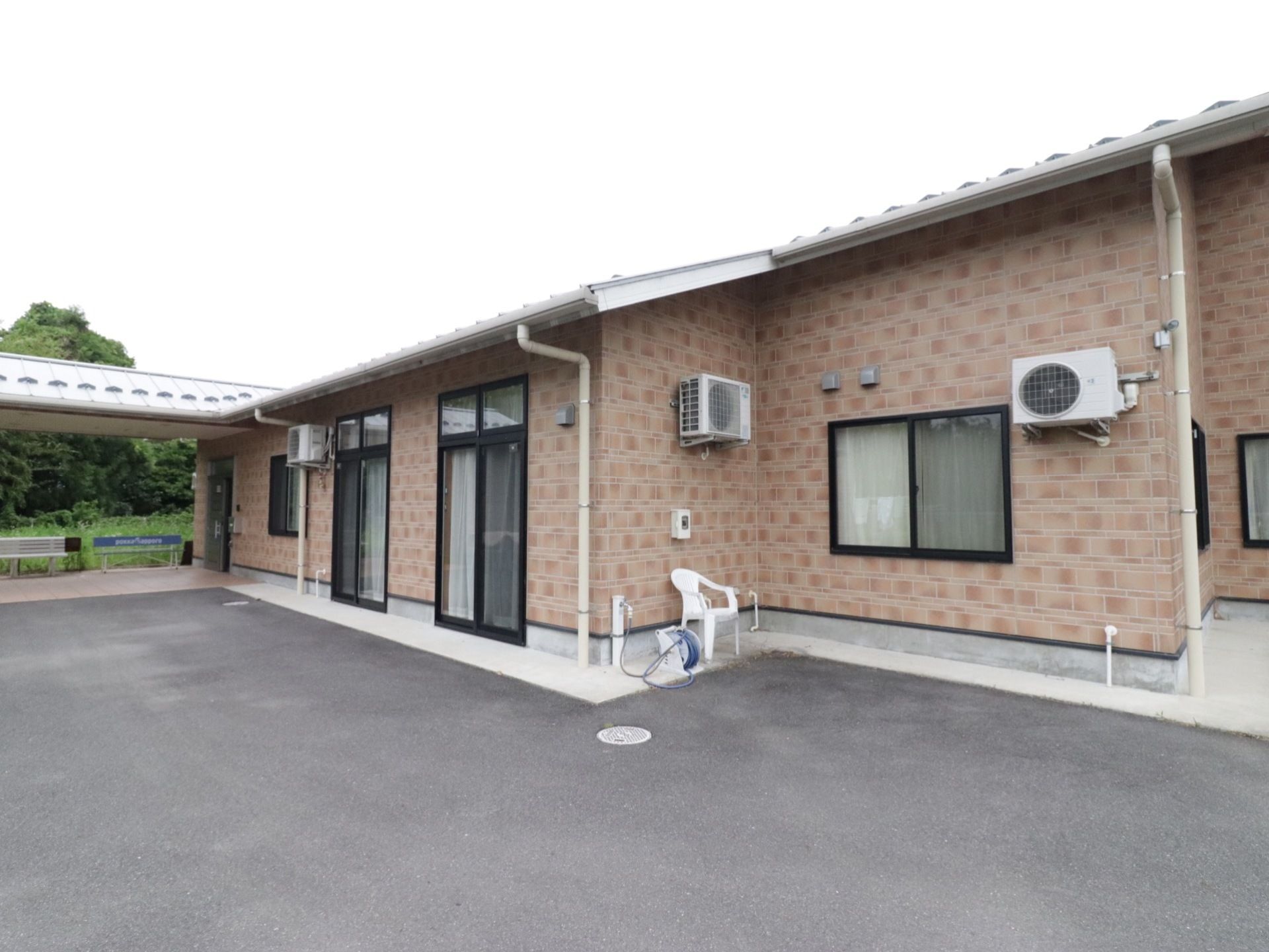 ブラウンの外壁の平屋建ての建物で、建物の前には広々としたスペースがある。周辺には緑が多く落ち着いた環境である。