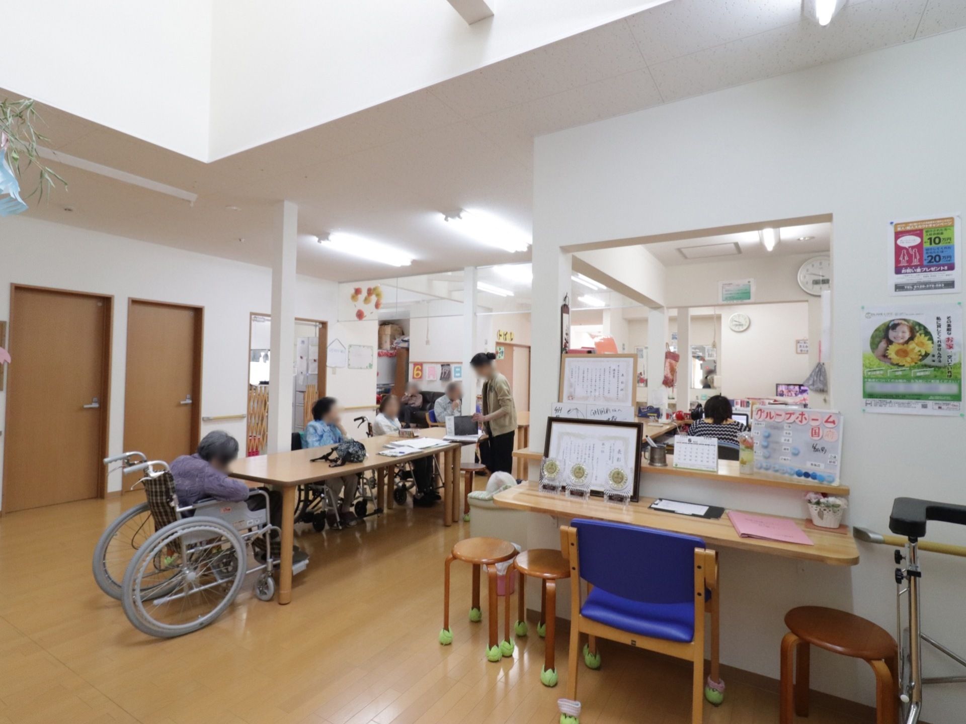 ゆったりとしたスペースに細長いテーブルが並んでいる。入居者様が椅子や車椅子に座っていて、スタッフが立って作業をしている。