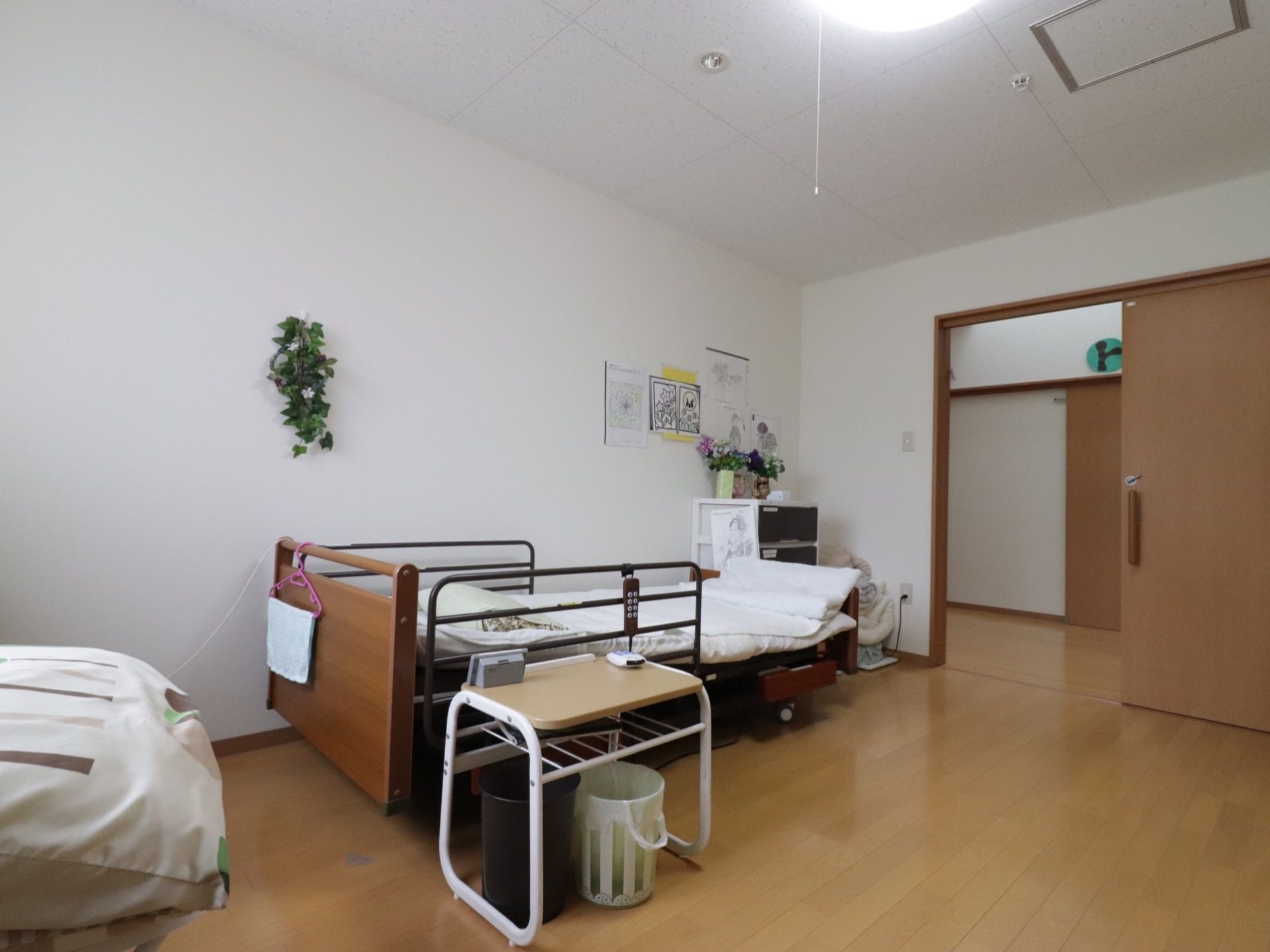 居室はゆったり広々としていてベッドや棚、ゴミ箱などが置かれている。壁には花やイラストなどが飾られている。