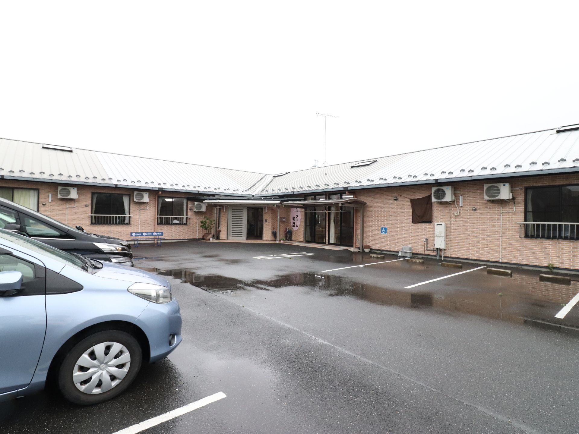 施設は平屋建てで、レンガ風の茶色い外壁で囲まれている。屋根はトタン葺き、手前の駐車場はアスファルトでできている。