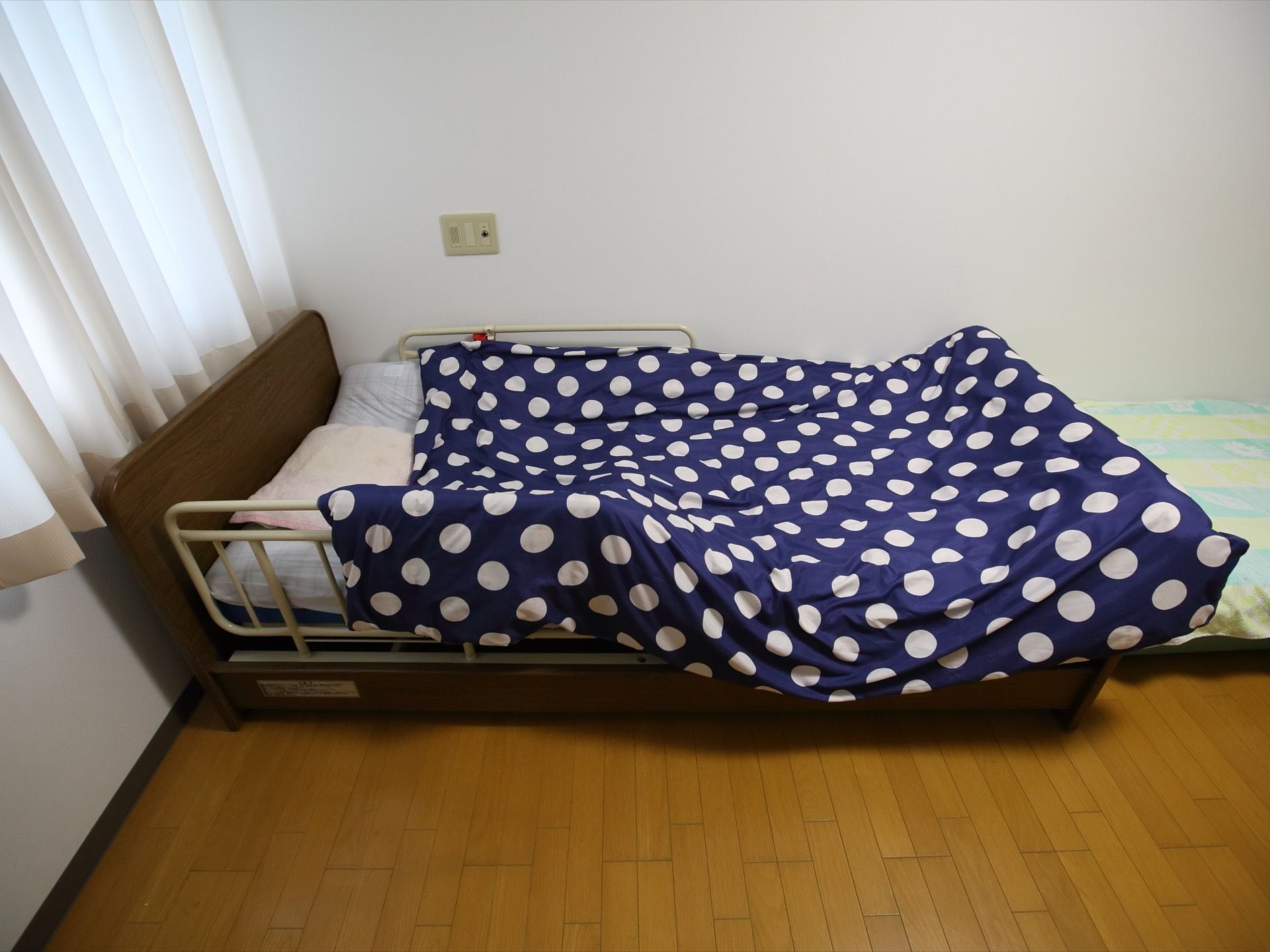 居室の窓近くに、介護用のベッドが１台置かれている。ベッドの近くの壁にはナースコールの差し込み口がある。