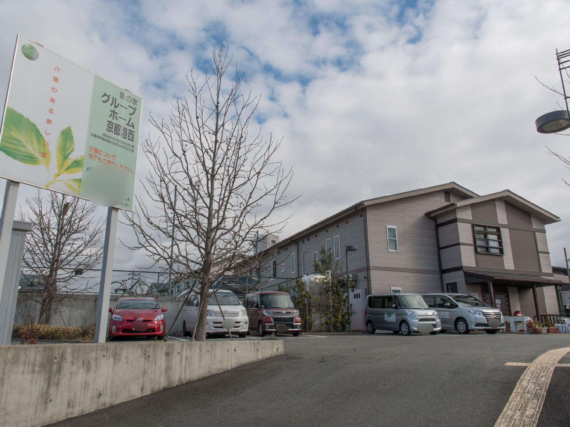 斜面に建っている施設で、全面に駐車場が設けてある。斜面手前に施設の名前が書いた看板が立っている。街路樹が点在している。