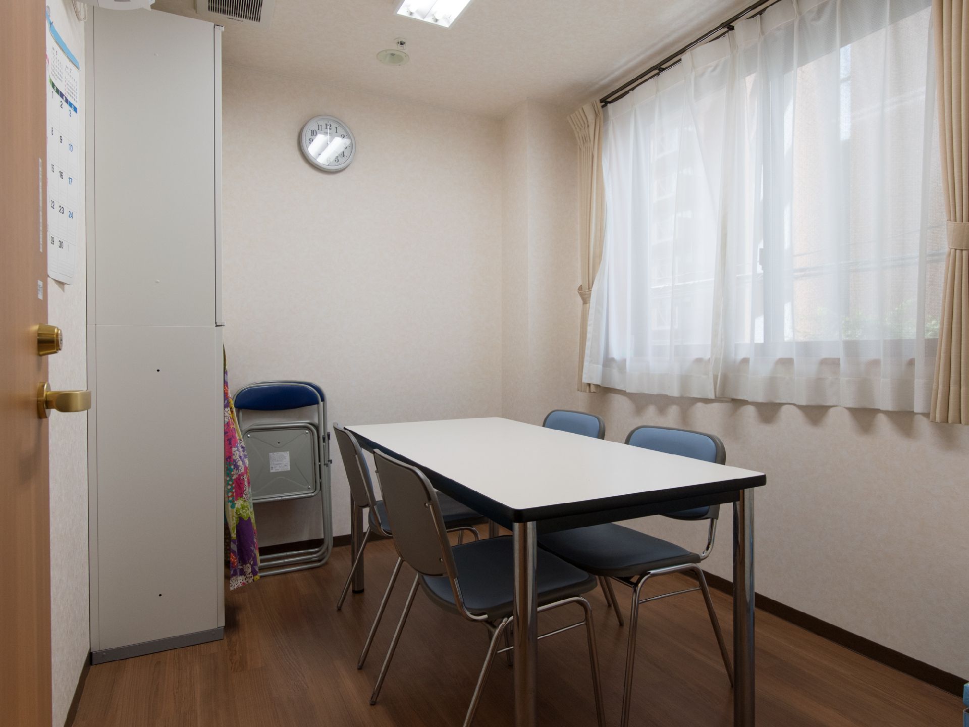 白い壁、茶色のフローリング、窓で構成された部屋に、机とパイプ椅子が置いてある。壁には壁掛け時計が飾ってある。