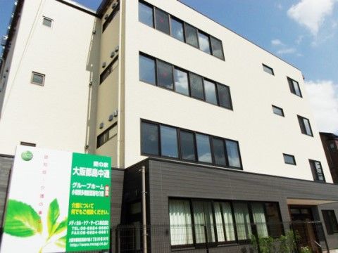 白い建物に、１階のロビー部分がダークブラウンに塗られている。施設前には緑色が映える大きな看板が立てられている。