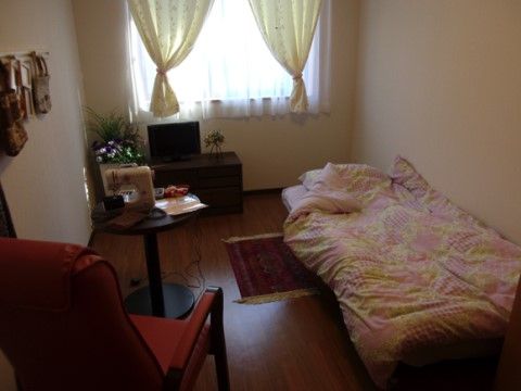 居室内の壁際にベッドが置かれている。窓際にテレビ台があり、小型の液晶テレビが乗せられている。１人用のソファセットがある。