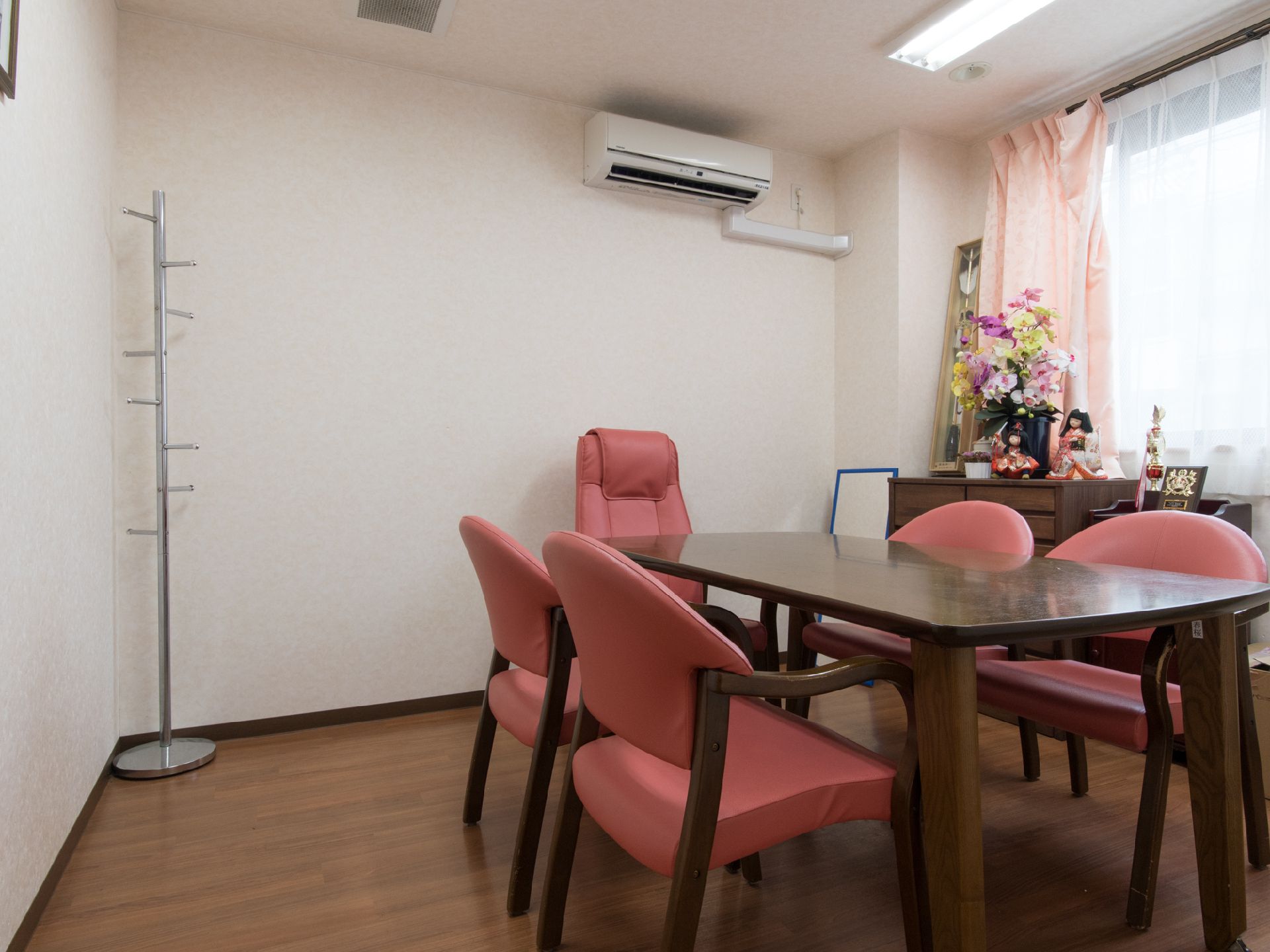 白い壁、ピンクのカーテンがかけてある窓、エアコンのある部屋で、ポールハンガーがコーナーにおいてある。