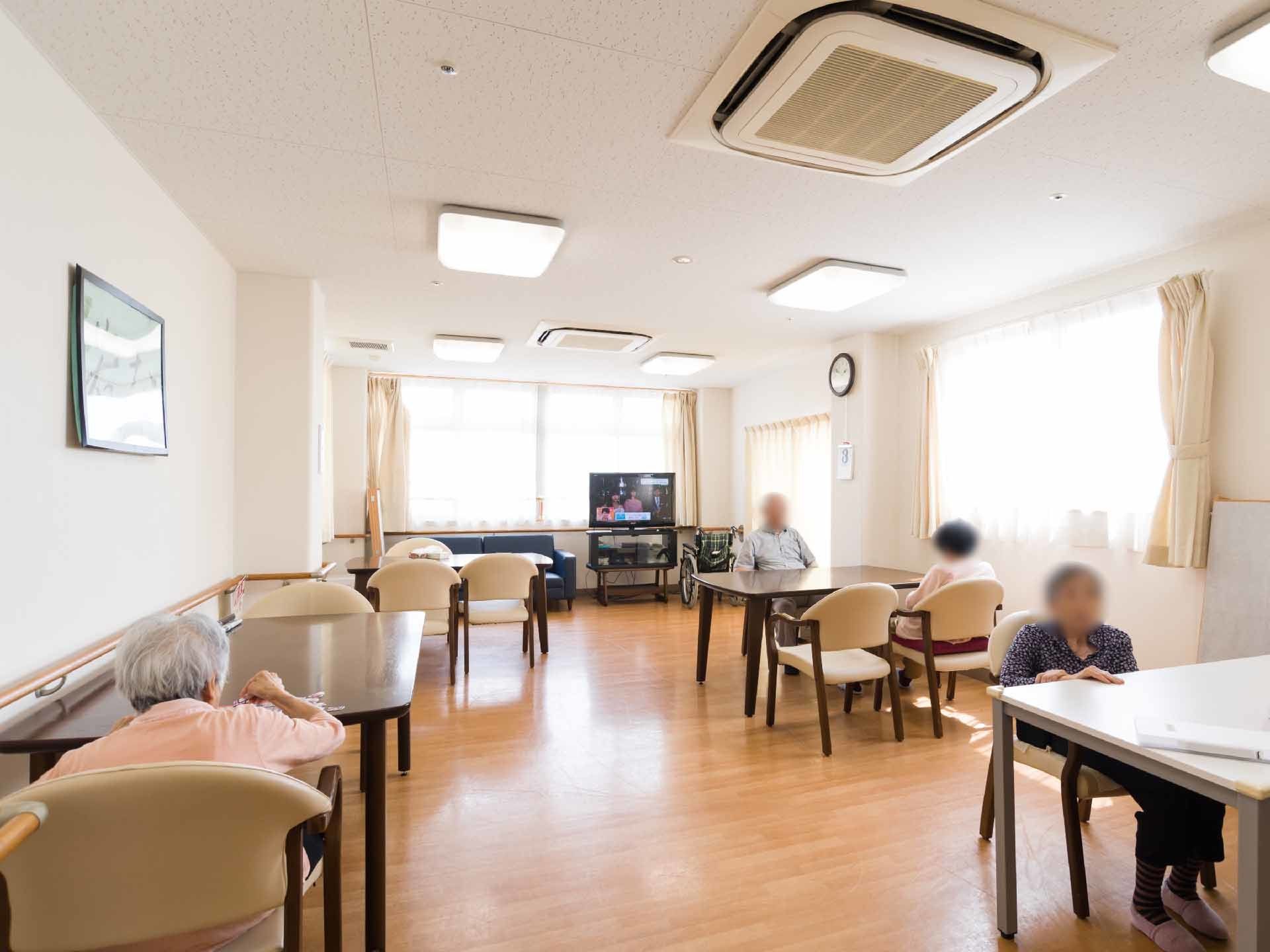 白い壁でフローリングのダイニングルームには、カーテンが取り付けてある窓がある。入居者が数名食堂にいる。