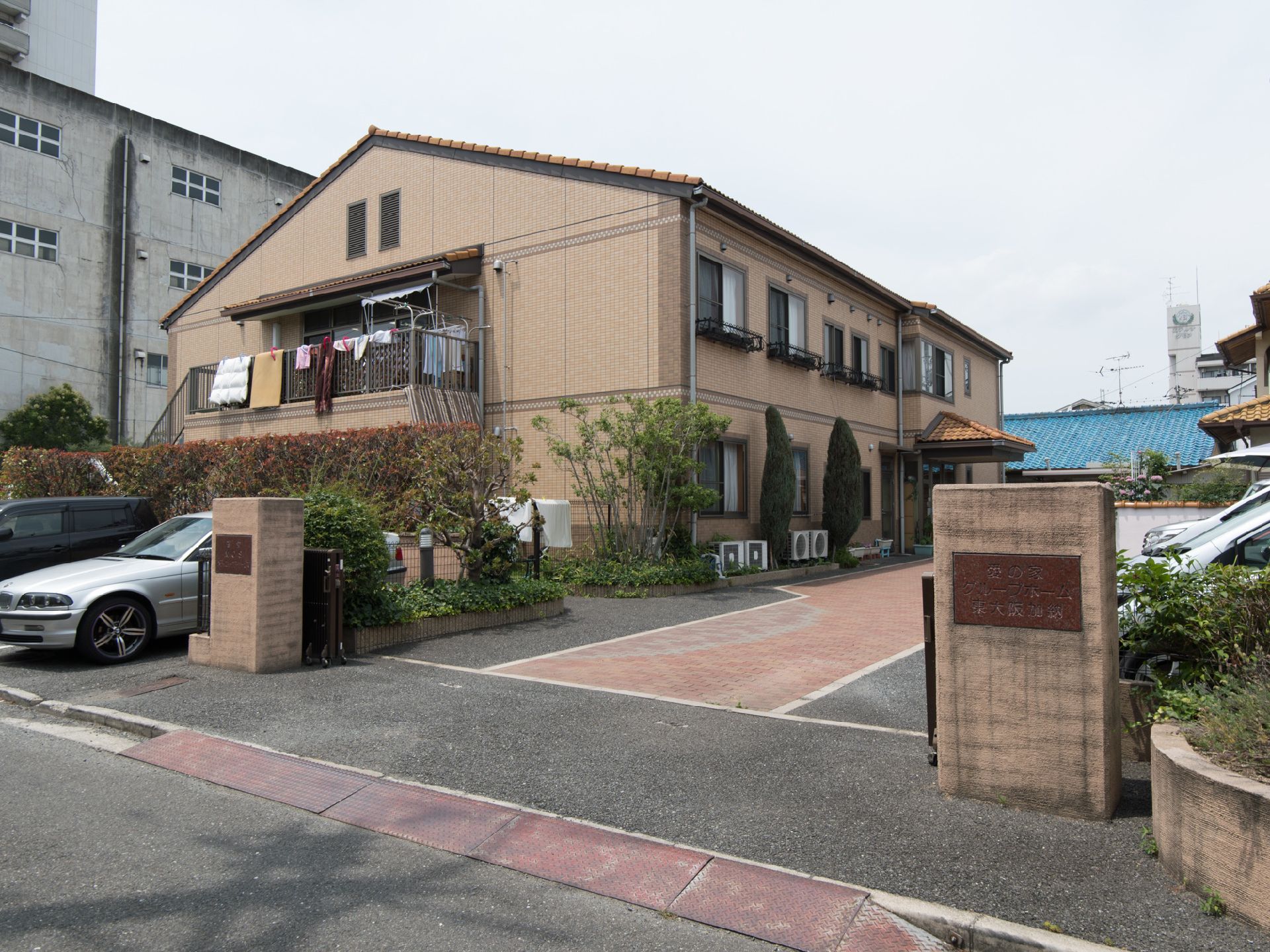 施設の周囲には植栽がしてある。室外機が数台置いてある。外壁が茶色でレンガぽくなっている。玄関の前にスタンドポストがある。