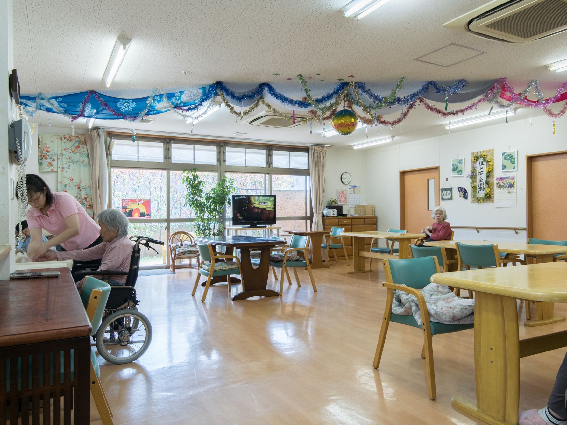 食堂で車いすの女性が手を洗うのを、職員がお手伝いしている。ダイニングテーブルにひじ掛けのついた椅子が置いてある。