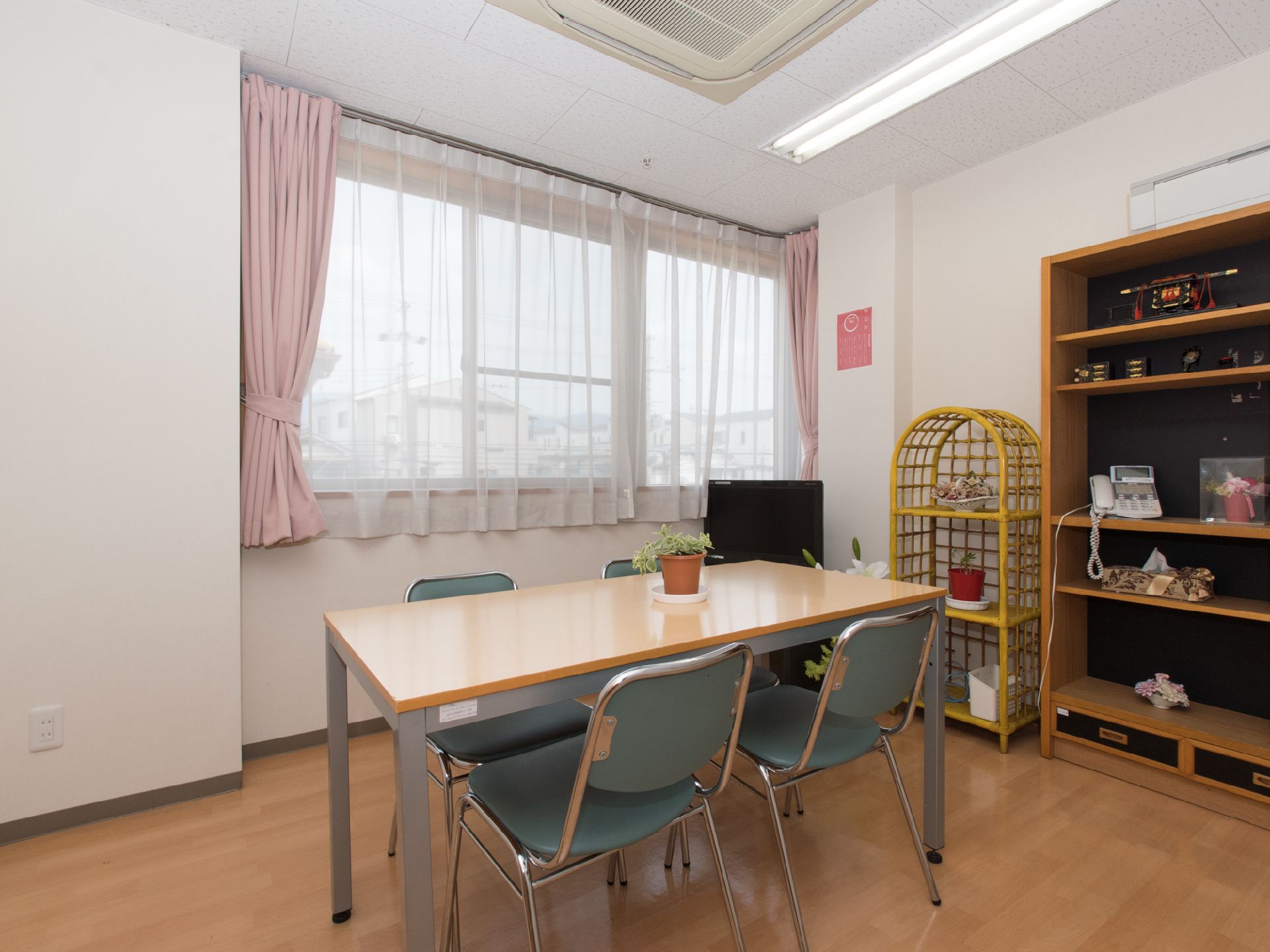 テレビ、棚などが置いてある部屋で、エアコンが完備である。観葉植物がデスク上にあるほか、棚にもある。パイ部椅子がある。
