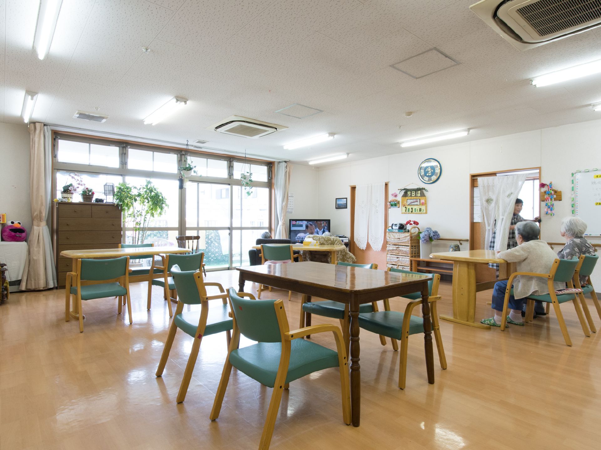 天井からフラワーバスケットが吊り下がっている。テレビの前に椅子を置いて干渉をしている入居者がいある。ドアには、暖簾がかかっている。