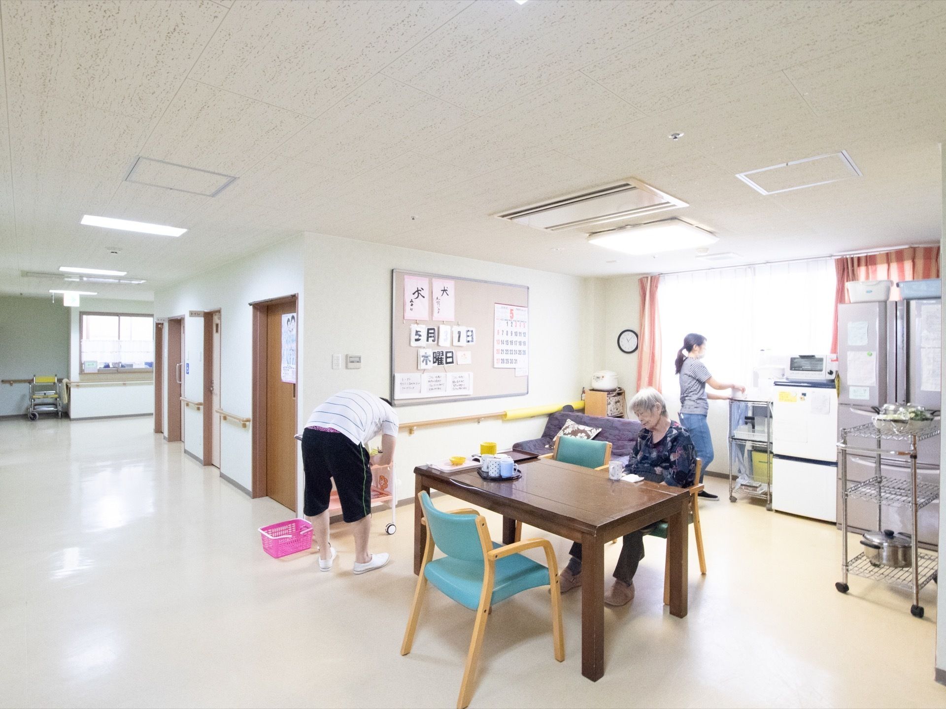 2つあるうちの1つの冷蔵庫の上にはトースターが置かれており、横では女性スタッフが作業をしている。テーブル席には入居者様座っている。
