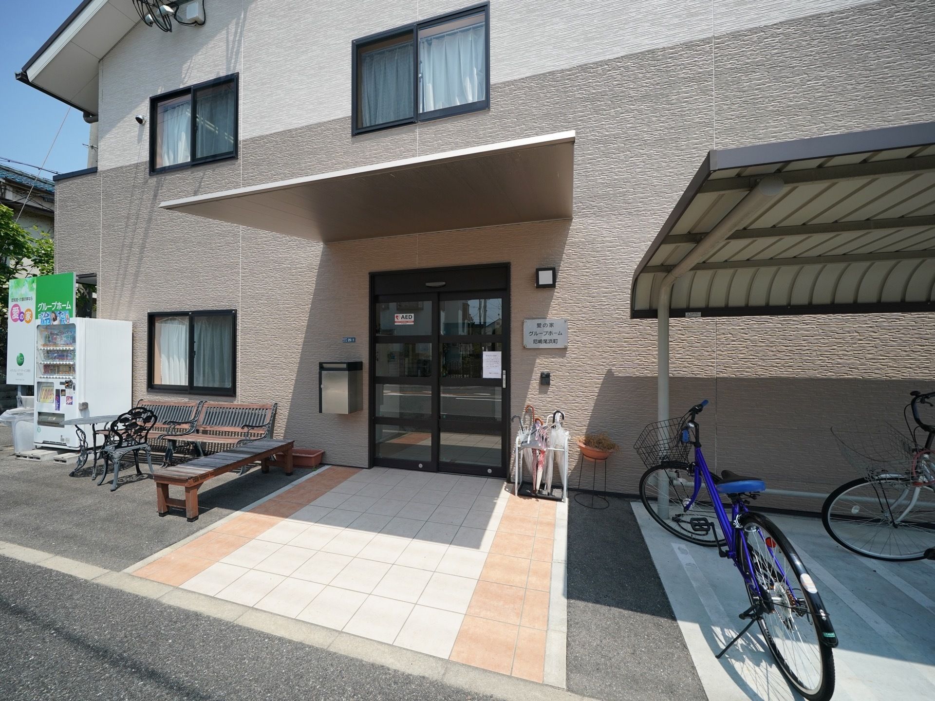 玄関前にオレンジとベージュのタイルが敷かれている。段差がなくフラットである。自動販売機のとなりには、ベンチが設置されている。