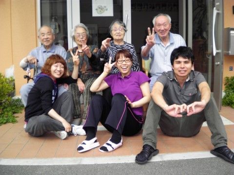 玄関前に集合して、笑顔を浮かべたりピースサインをしたりしている入居者様たちとスタッフ
