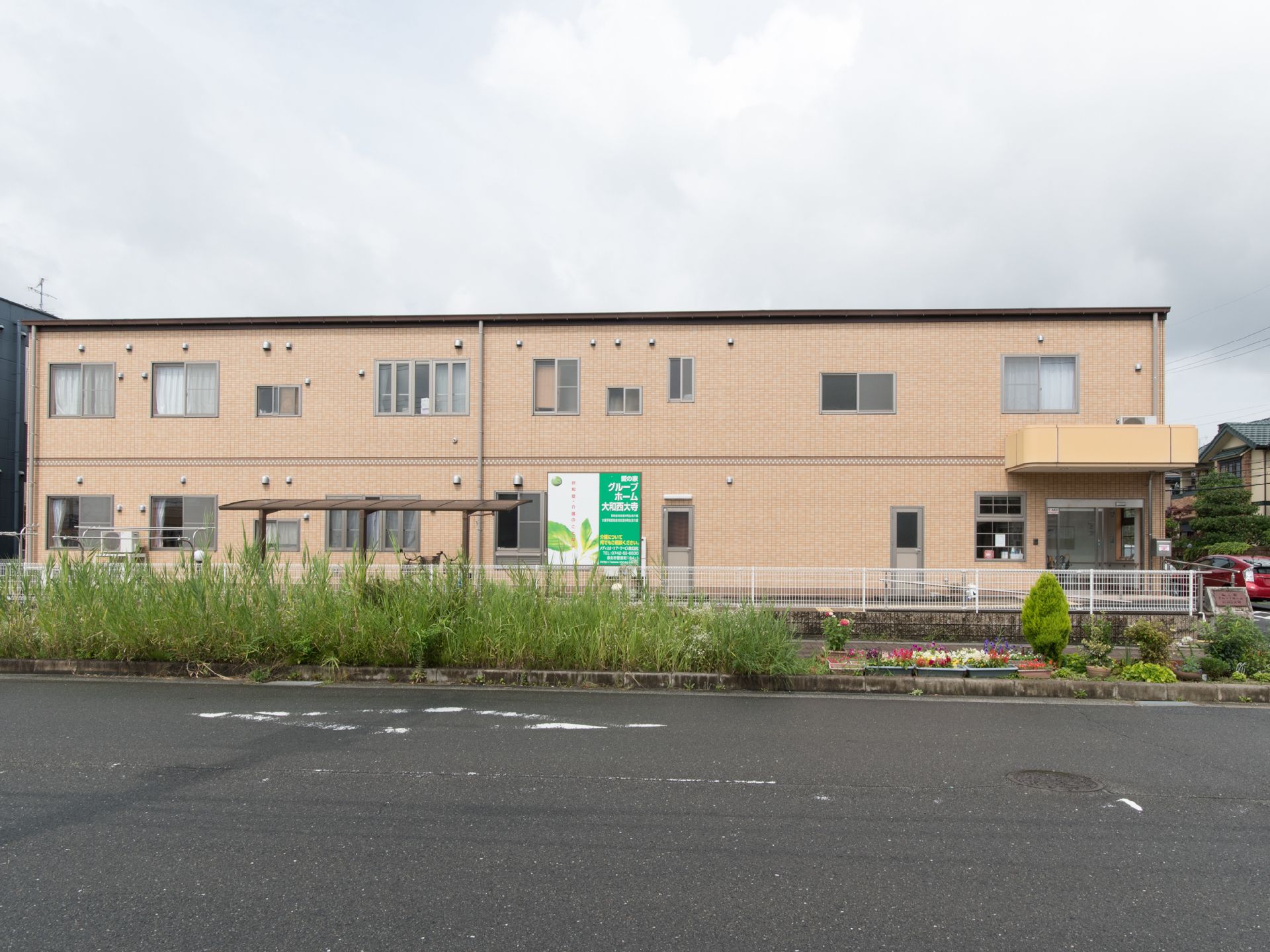 茶色の外観で、白いフェンスで囲ってある。歩道と道路の境目にたくさんのプランターがある。スロープそばに駐車場がある。