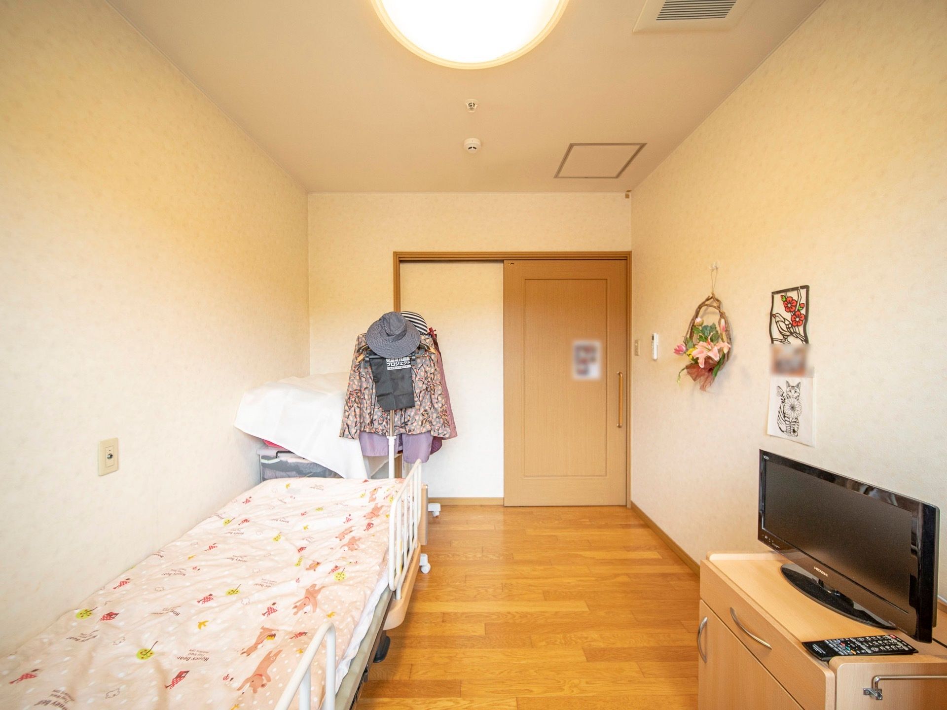 木目調の床の居室である。介護用ベッドがあり、低い棚の上にはテレビが置かれている。ハンガーラックとアクリルケースがある。