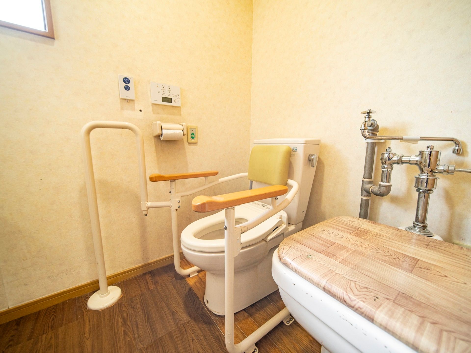 ダークブラウンの床の広いトイレである。便座には背もたれとひじ掛け、立体的な手すりが付いている。壁には小窓が付いている。