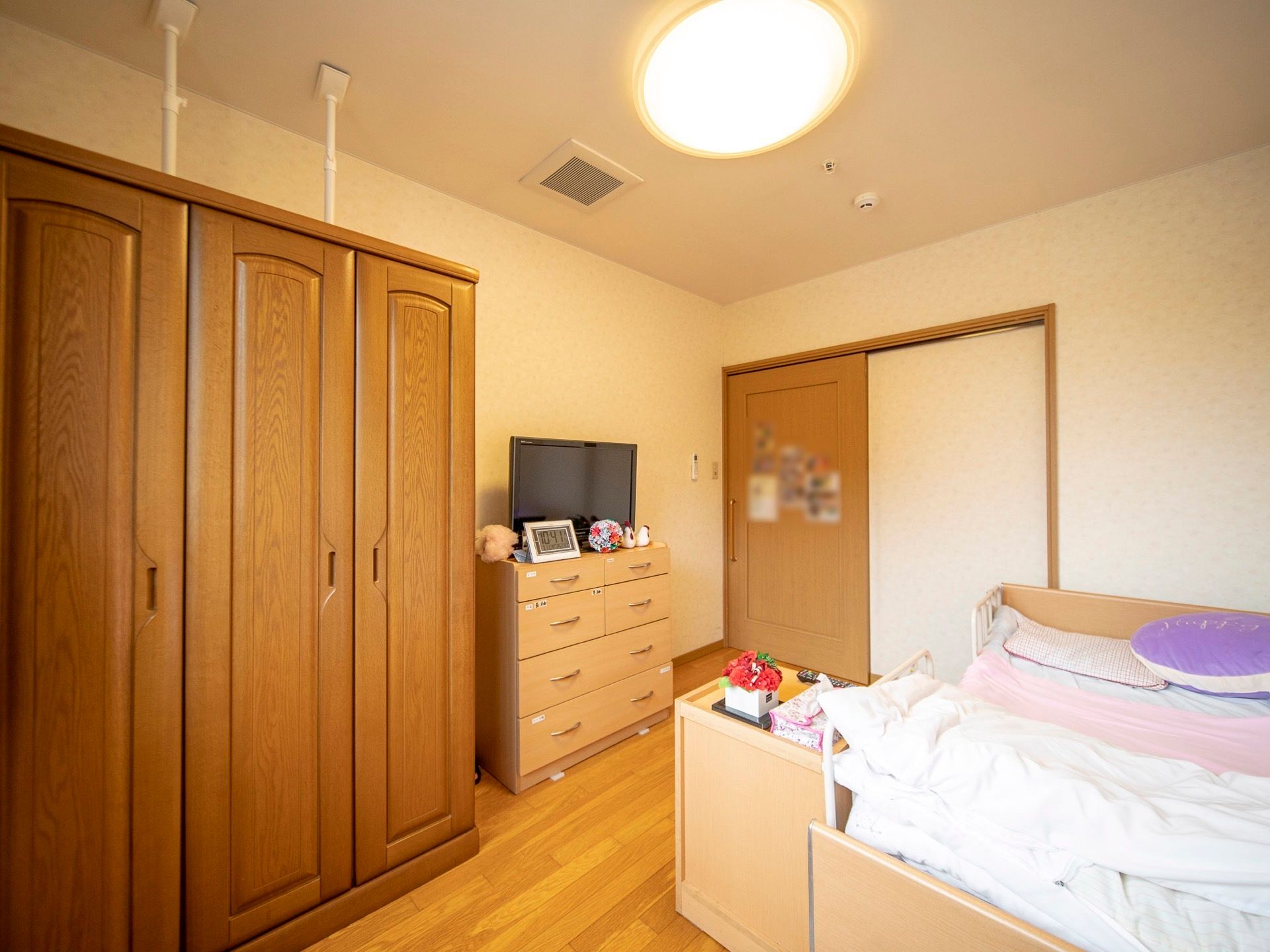 介護用ベッドが置かれている居室である。大きなグローゼット型のタンスと、背の低いタンスがあり、テレビが置かれている。