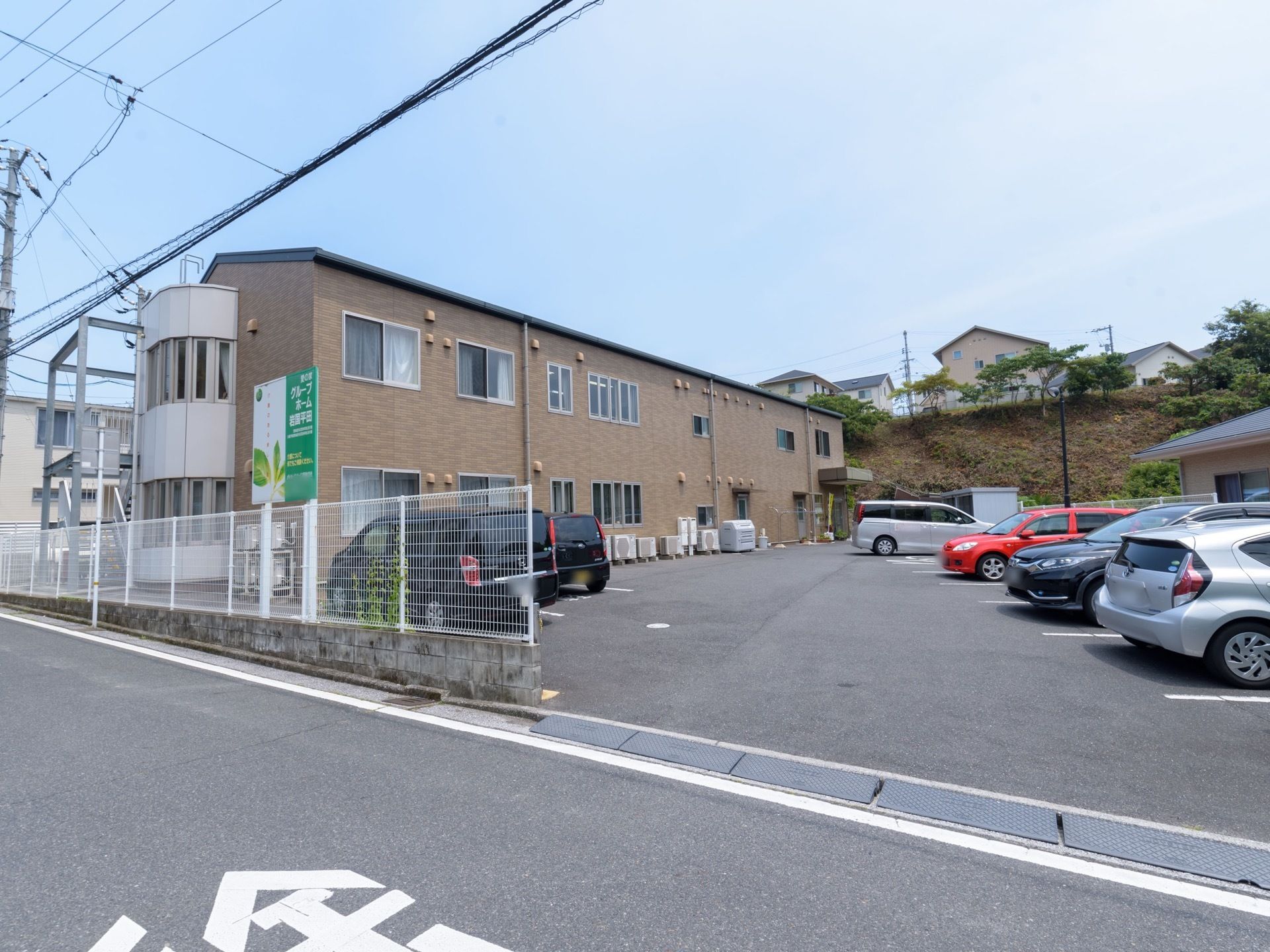 施設は2階建てで、ベージュ色のレンガ調の外観である。駐車スペースの近くにはフェンスがあり、施設の看板が立てかけられている。