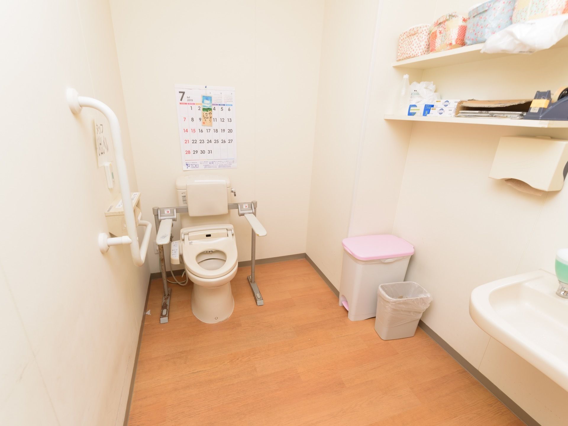 広々としたトイレには洋式便器が設置されている。トイレタンクの後ろの壁にはカレンダーが貼られている。ゴミ箱が置かれている。
