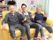 黄色いソファーに男性が３人座って手を振っている。手前にあるテーブルには置物が並んでいる。奥に加湿器などが置かれている。