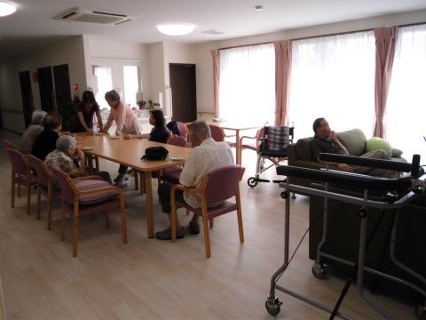 食堂に集まって、立っている２名のスタッフの方を見ている入居者様たち