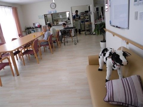 木目調のフローリングの部屋である。左側に木製のテーブルが並び、揃いの椅子が置かれている。数人の入居者様が座っている。