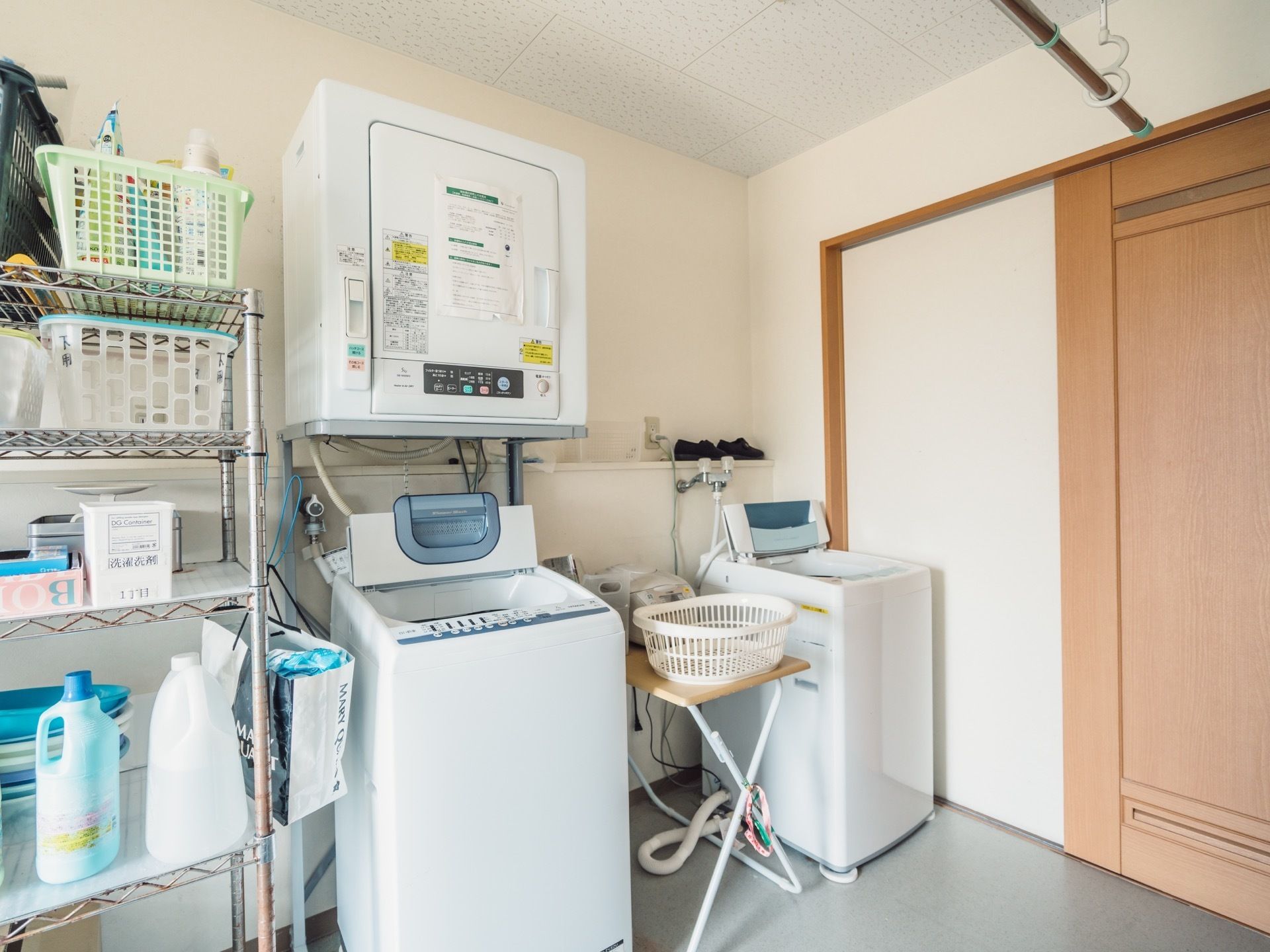 縦型式の洗濯機が２台と乾燥機が１台並んでいる。棚には洗剤や掃除用スプレーがきれいに整頓されて置かれている。