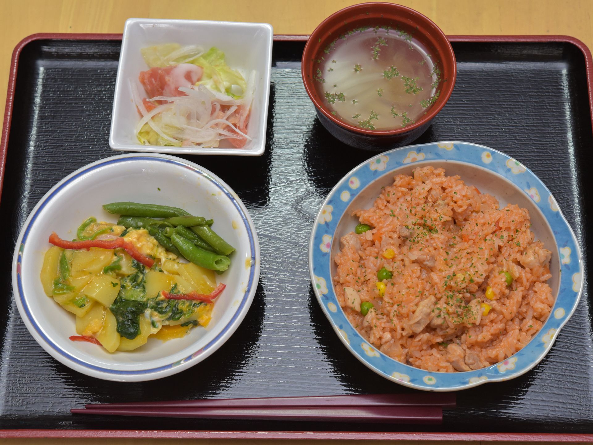 黒色のトレーの上にチキンライスと汁物とメインおかずに小鉢が1つ置かれている。栄養バランスと彩りと品数をトータル的に考えられた献立である。