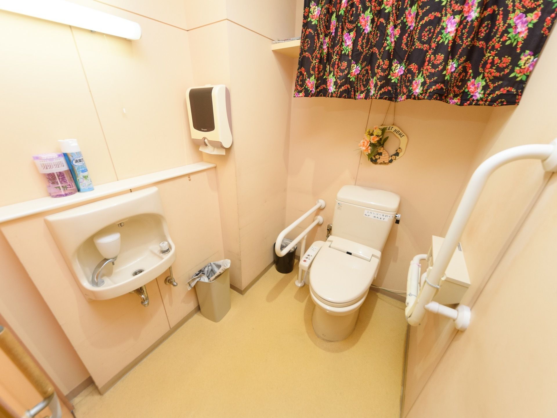 洗面台が備わっている広いトイレである。壁には、可動式やＬ型手すりが取り付けられており、備品が置ける棚が設置されている。