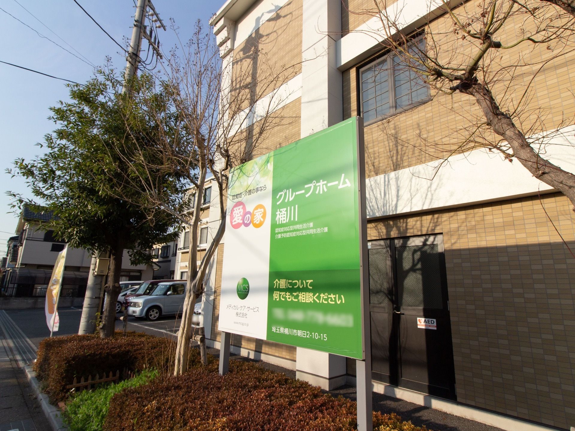 植え込みの中に看板が立っており、施設名が書かれている。まわりには木が植えてあり、後ろには茶色の外壁の建物がある。