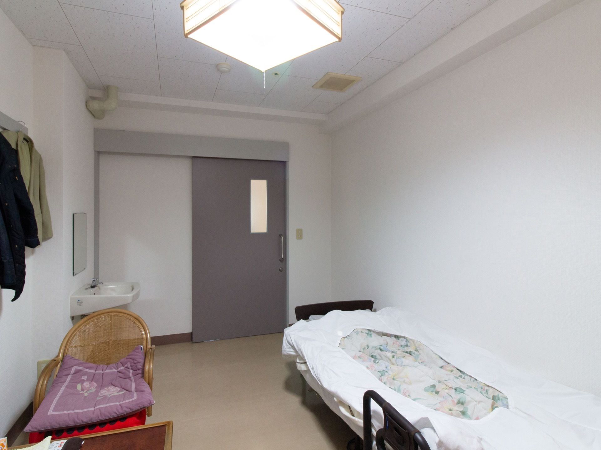 ドアはグレーの大きな引き戸になっていて、左側には洗面台と鏡が設置されている。部屋にはベッドや椅子などが置かれている。