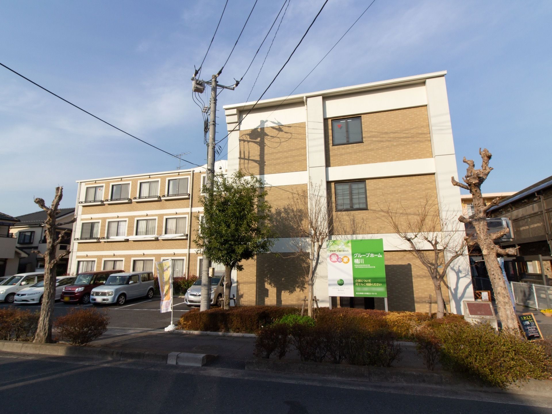 施設は３階建ての茶色い建物になっていて、左側には駐車場がある。建物の前の歩道には植え込みや木がある。