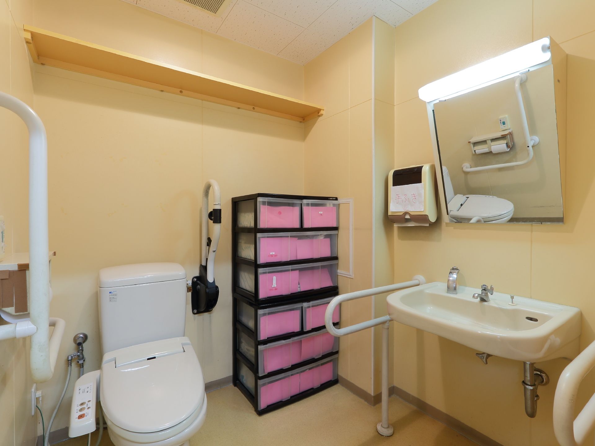 クリーム色の壁の内装で、洗面台が設置されたトイレである。手すりが設置され、便器上の方には黄色の棚がある。