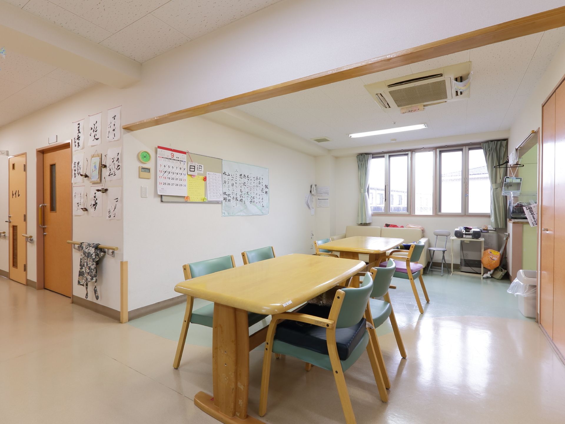廊下に面する共有スペースである。木製のテーブルと椅子が並べられている。横一面は窓になり、壁にはカレンダーなどが貼ってある。